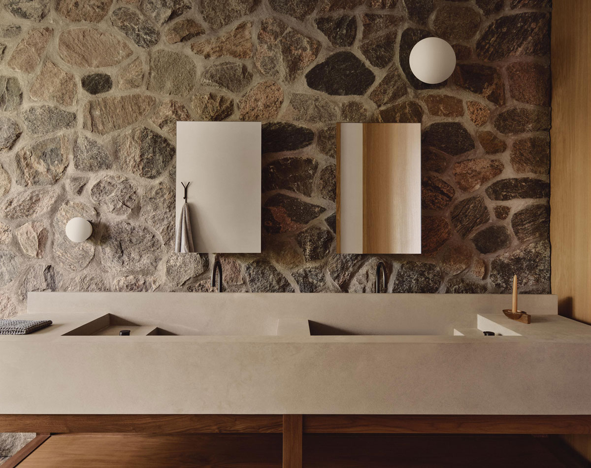 Double-basin stone vanity against a fieldstone wall, with brass faucets, rectangular mirrors, and globe lights.