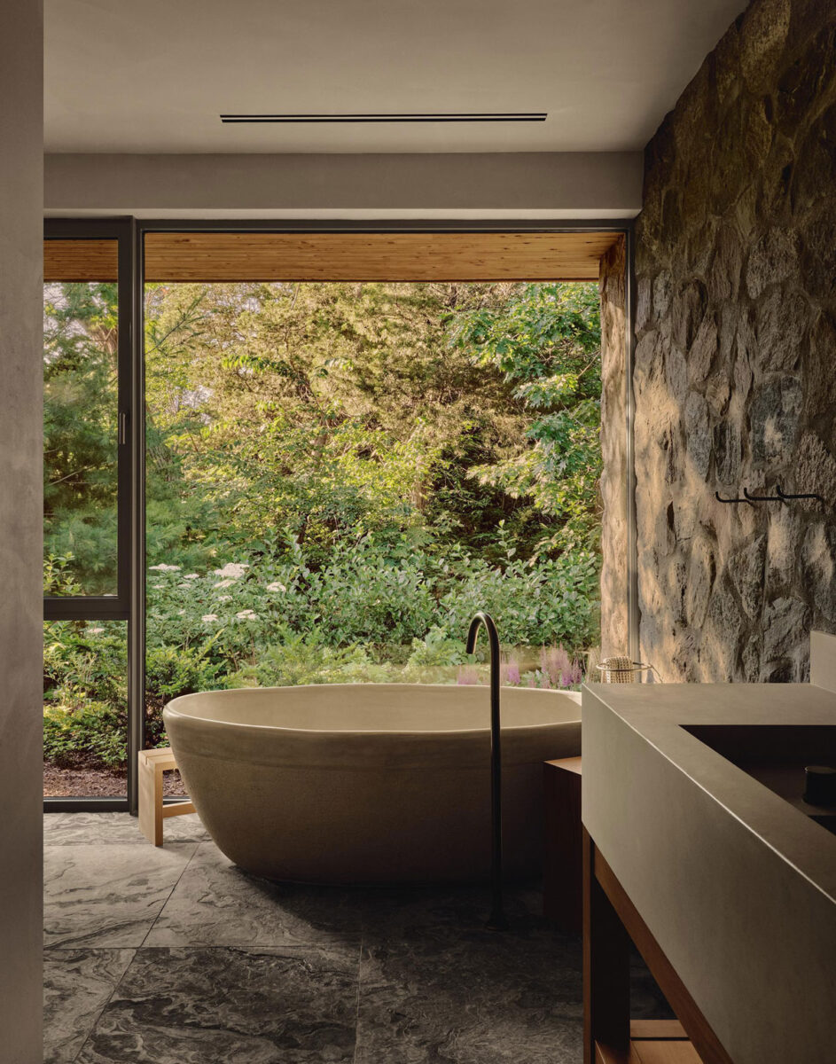 Freestanding ceramic bathtub beside a fieldstone wall and a large window overlooking woodland.
