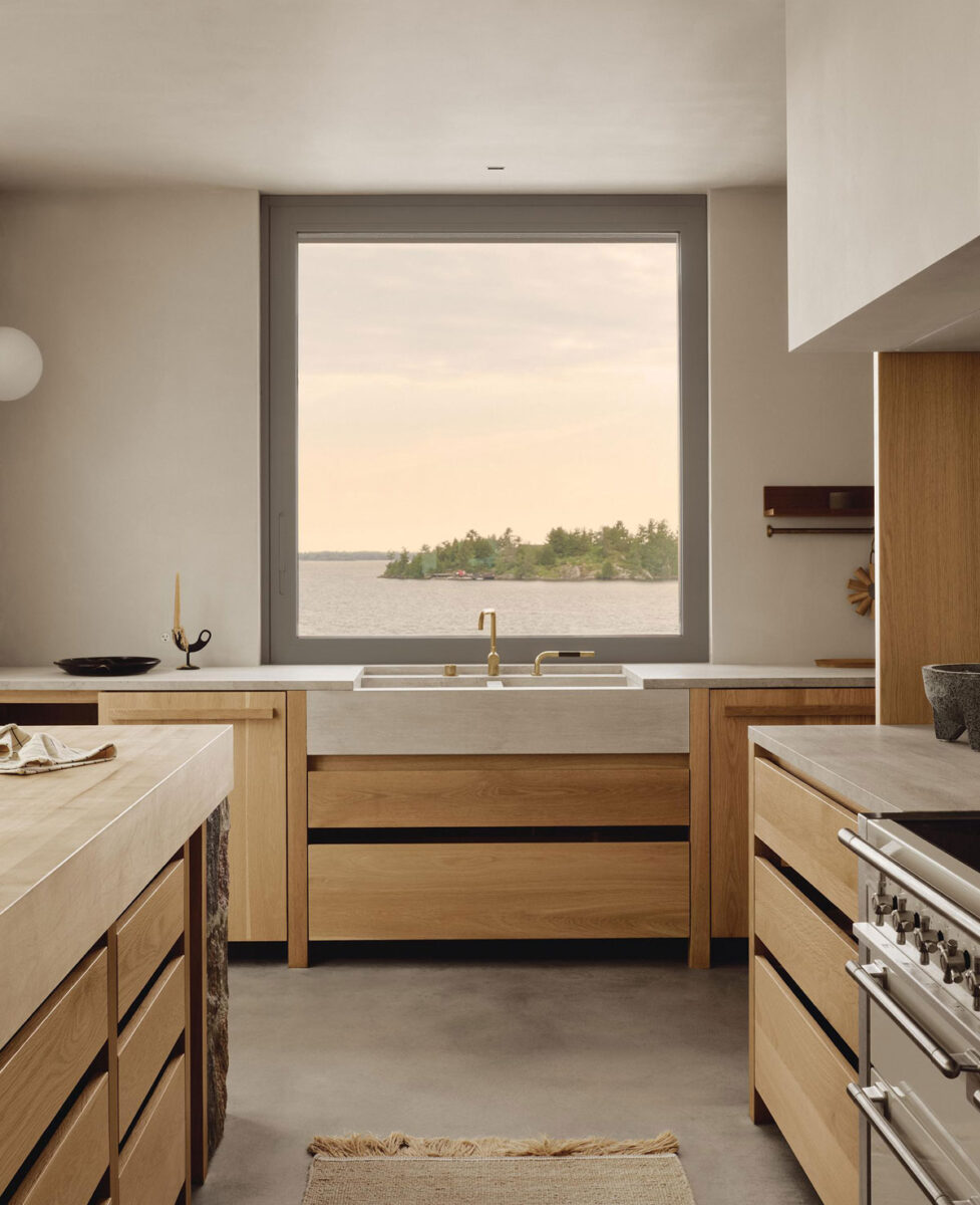 Oak kitchen cabinetry and a stone countertop beneath a window overlooking Georgian Bay at sunset.