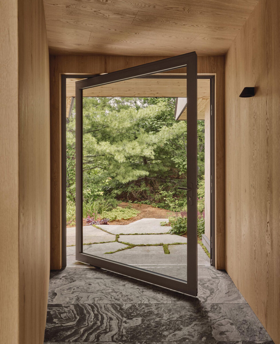 Large pivot glass door in an oak-lined passage, connecting textured stone flooring to a garden path.
